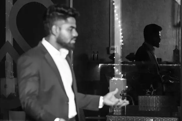 Black and white frame of the speaker gesturing, a figure watching behind him
